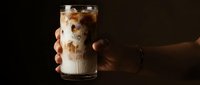 Hand holding a glass of iced coffee with cream swirl against dark background, perfect for beverage, cafe, and lifestyle marketing materials.