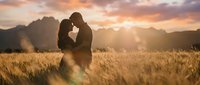 Romantic couple silhouette embracing in golden wheat field at sunset with dramatic mountain landscape and warm sunlight creating intimate moment for love concepts.