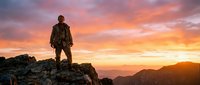 Male hiker standing on rocky mountain peak at sunset, wearing outdoor gear, gazing at golden sky and distant valleys below for adventure and travel concepts.