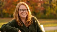 Happy blonde woman with glasses wearing green sweater smiling outdoors in autumn park with colorful fall foliage background for lifestyle content.
