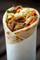 Grilled chicken wrap filled with fresh vegetables, shredded cabbage and carrots in tortilla bread, close up view of street food on dark background.
