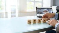 Man in business attire arranging wooden blocks spelling tax with percentage symbol at desk with laptop, representing tax planning and financial management for accounting and business purposes.