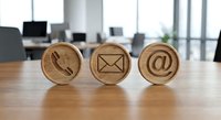 Wooden contact icons on office desk showing phone, email and at symbols for business communication and customer service concepts.