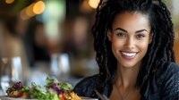 Young African American woman with dreadlocks smiling at camera while enjoying healthy salad meal in modern restaurant setting with warm bokeh lighting.