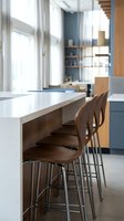 Modern kitchen island with white countertop and wooden bar stools, featuring contemporary design with natural light from windows and minimalist shelving in background.
