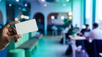 Closeup of hand holding blank white business card in modern office environment with blurred colleagues working in background, suitable for corporate branding and professional networking materials.