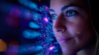 Young woman illuminated by futuristic digital technology interface with glowing particles and data visualization in blue and purple neon lights against dark background.