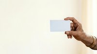 An older male hand with medium-dark skin tone holds a blank white business card horizontally against a neutral beige background, ideal for corporate identity, networking, and professional branding mockups.