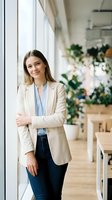 Confident young Caucasian businesswoman in beige blazer standing with crossed arms in modern bright office space with plants and natural light.