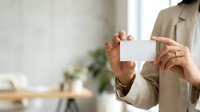 Unrecognizable male professional in beige blazer holding blank white card in modern office workspace with natural light and green plant decor.