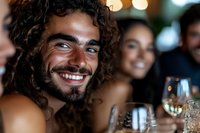 Young Mediterranean man with curly dark hair and beard smiling genuinely at dinner party with friends, enjoying wine and social gathering in evening setting.