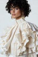 Young mixed race woman with curly black hair wearing cream ruffled high-neck blouse and ornate gold earrings against white background, fashion portrait.
