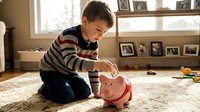 Young boy, approximately 6-8 years old, Caucasian ethnicity, placing coin into pink piggy bank while sitting on carpeted floor in home setting. Child wearing striped sweater engaged in savings and financial education activity.