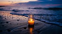Glowing candle in glass jar on sandy beach at sunset with ocean waves and dramatic purple sky creating peaceful romantic atmosphere.