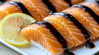 Fresh salmon nigiri sushi with teriyaki sauce drizzle and lemon slice garnish on white plate, close up view showing vibrant orange fish texture and glossy glaze.