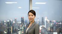 Asian businesswoman in gray blazer standing confidently before cityscape window, suitable for corporate, leadership, and professional business concepts.