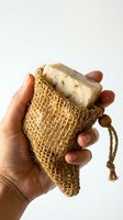 Hand holding a natural herbal bar soap in a burlap sisal exfoliating bag, eco-friendly zero waste bathroom accessory on white background.
