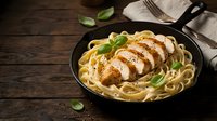 Sliced grilled chicken breast over creamy pasta in cast iron skillet with fresh basil on rustic wooden table for restaurant menu and cooking content.