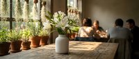 White flowers in ceramic vase on wooden table with potted plants by sunny window in cozy restaurant with diners enjoying meal in warm natural light.