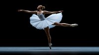 Young Caucasian ballerina in white tutu performing arabesque on pointe against dark background, graceful dance pose showing strength and elegance in classical ballet.