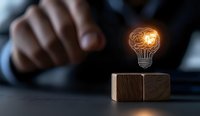 Glowing brain lightbulb hologram floating over wooden cube with hand in background, representing innovation and creative thinking in business concept.