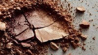 Broken eyeshadow palette with shimmering bronze and pink tones surrounded by loose powder, showing makeup texture and damage.