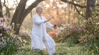 Senior Caucasian woman practicing tai chi in peaceful garden setting with blooming flowers during golden hour sunrise meditation and wellness routine.