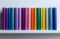 Row of colorful hardcover books standing in line on white shelf against white wall, creating rainbow gradient pattern from purple through pink to yellow and blue.