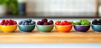 Colorful ceramic bowls with assorted fresh berries and fruits arranged in rainbow order on wooden surface, perfect for healthy eating concept.