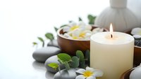 Spa wellness setup with burning candle, plumeria flowers in wooden bowl, massage stones and eucalyptus leaves for relaxation and aromatherapy treatments.