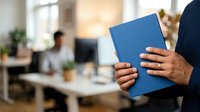 Cropped view of professional holding blue notebook in modern office workspace with colleagues working at desks in background, suitable for business, corporate training, and workplace concepts.