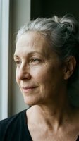 Senior woman with gray hair in bun, fair complexion, wearing black shirt, gazing thoughtfully by window with natural light. Portrait for aging, wellness, lifestyle content.