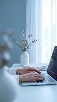 Female hands typing on laptop keyboard at modern white desk with eucalyptus branch in vase by window for remote work and productivity concepts.