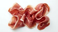 Thinly sliced prosciutto ham with marbled texture arranged in elegant curls on white background, perfect for gourmet cooking and culinary presentations.
