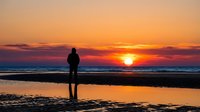 Surfer in wetsuit standing on beach at golden sunset, holding surfboard, watching ocean waves during dusk. Coastal lifestyle.