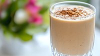 Creamy cappuccino or latte beverage topped with cocoa powder in clear glass with blurred flowers and foliage background.