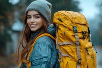 Young Caucasian woman hiker with bright yellow backpack smiling in rain, wearing gray beanie and waterproof jacket. Adventure travel lifestyle outdoors in nature.