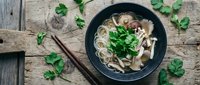 Asian noodle soup with mushrooms and fresh herbs in black bowl on rustic wooden table with chopsticks for healthy comfort food recipes.