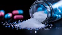 White crystalline powder spilling from glass bottle with pink and blue capsule pills scattered on dark surface in dramatic lighting, medical concept.
