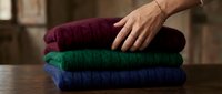 Hand placing folded colorful knit sweaters in burgundy, green and blue on wooden surface. Cozy winter clothing stack for seasonal wardrobe organization.