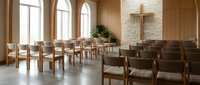 Modern chapel interior with wooden chairs arranged in rows facing a wooden cross mounted on a light stone wall, large arched windows providing natural light, potted green plant, and minimalist design for worship spaces and religious ceremonies.