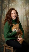 Happy young Caucasian woman with curly red hair holding red fox in green sweater against autumn forest background for wildlife conservation campaigns.