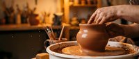 Skilled potter's hands shaping clay vessel on spinning wheel in traditional ceramic workshop with warm lighting and artisan tools in background.
