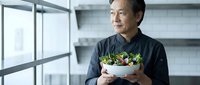 Middle-aged Asian man in black chef's coat holding fresh salad bowl in modern kitchen with shelving. Healthy eating and culinary concept.