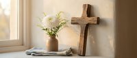 Rustic wooden cross with white flowers in ceramic vase on windowsill creating peaceful spiritual home decor atmosphere for meditation and prayer space.