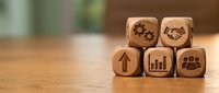 Wooden cubes with business icons including gears, handshake, arrow, chart and people symbols on desk for corporate strategy and teamwork concepts.