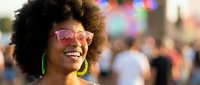 Happy young African American woman with afro hairstyle wearing colorful sunglasses and green earrings smiling at outdoor festival or street event.