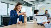 Professional businesswoman building wooden letter blocks spelling AGILE in modern office meeting room with colleagues working on laptops in background.