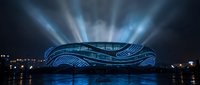 Futuristic stadium with flowing architecture illuminated by dramatic light beams at night, reflecting on water surface creating stunning architectural photography.