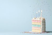Slice of pastel rainbow layer cake with white frosting and lit birthday candles on light blue background, scattered with colorful sprinkles. Celebration dessert.
